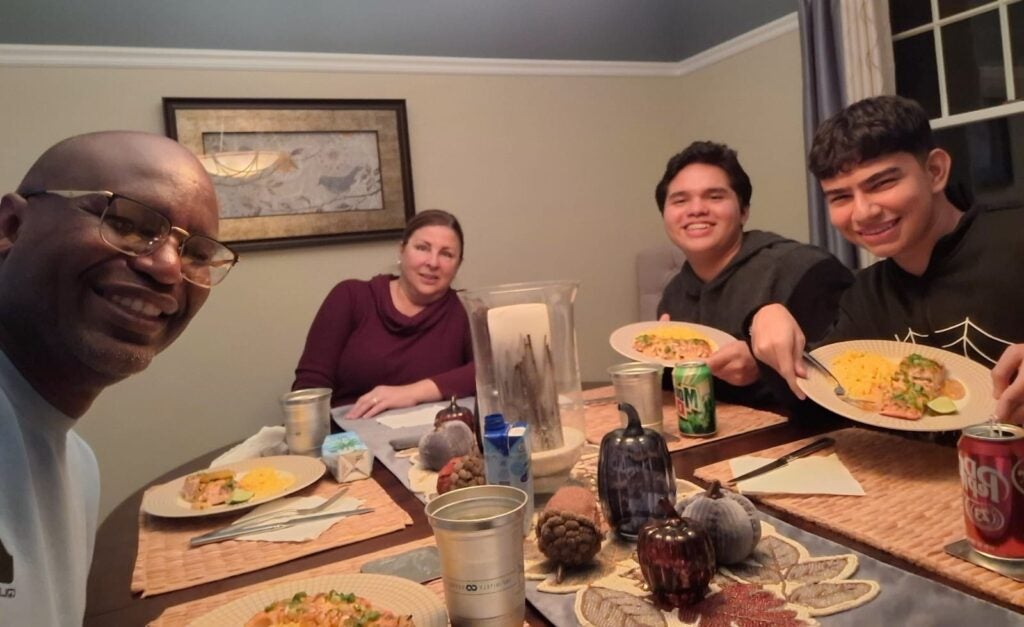 Josimar Madrigal seated at a dining table sharing a meal and conversation with his host parents, Mr. Garvin and his host mother, in Cleveland.